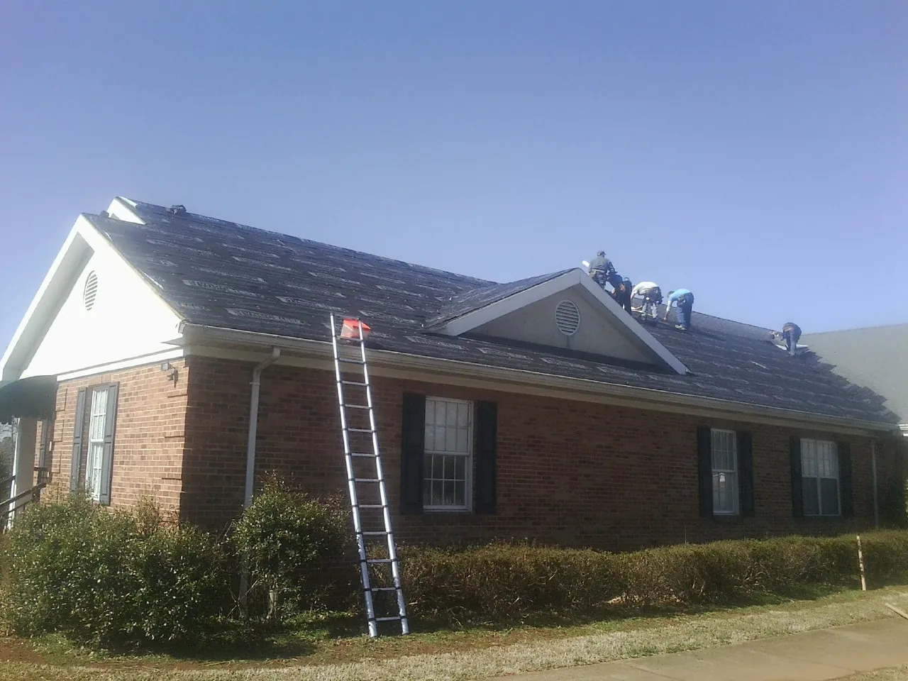 Crew tearing shingles off a brick ranch roof with a ladder set — Emergency Roof Repair work by our crew in King George