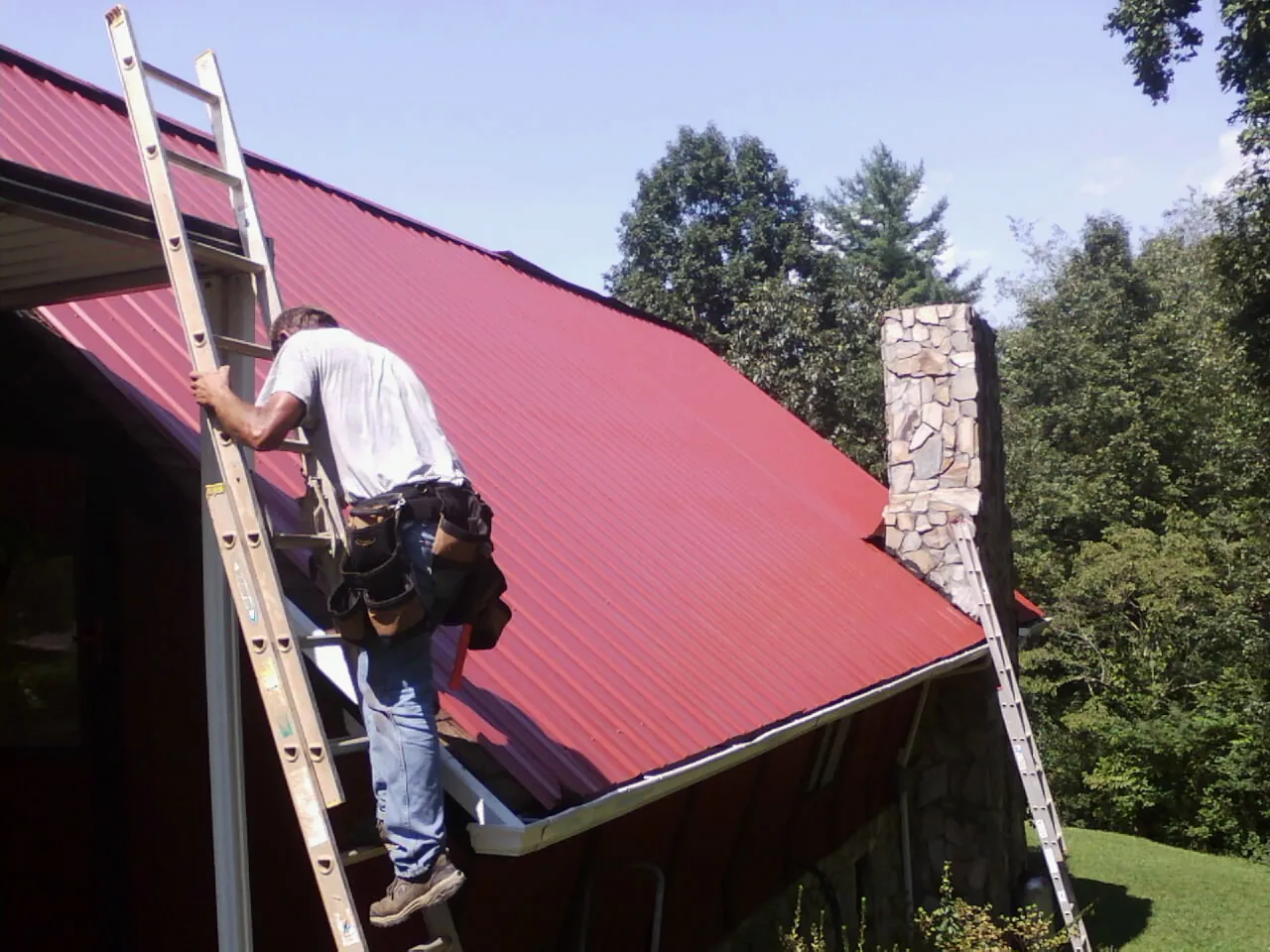 Roofer on a ladder beside a red metal roof and stone chimney — Soffit & Fascia Repair by Roofing Brothers in King George